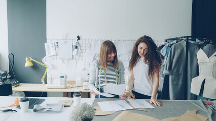 Wall Mural - Young woman clothing desigher is drawing sketch when her coworker is coming to her with garment draft, showing and improving image and discussing it with colleague.