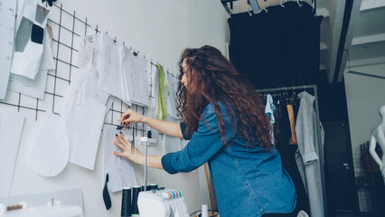 Wall Mural - Low angle view of young attractive fashion designer looking at sketches and hanging drawings on wall in modern loft style studio. Large collection of illustration above tailoring desk.