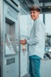 © Migue Suarez/Wirestock Creators - Smiling young Spanish male in front of an ATM machine, vertical shot