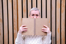 Woman Hiding Face Behind Open Book In Front Of Wall