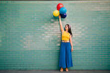 Woman With Hand Raised Holding Multi Colored Balloons In Front Of Teal Brick Wall