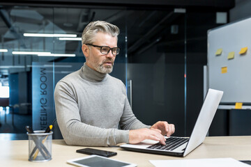 Senior and experienced programmer works in modern office at laptop, male web designer developer in glasses writes code programs new application, mature businessman works at desk.