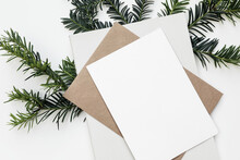 Festive Closeup Of Christmas Blank Greeting Card, Invitation Mockup Template. Craft Envelope, Diary And Green Yew Tree Branch Isolated On White Table Background. Winter Stationery. Flat Lay, Top View.