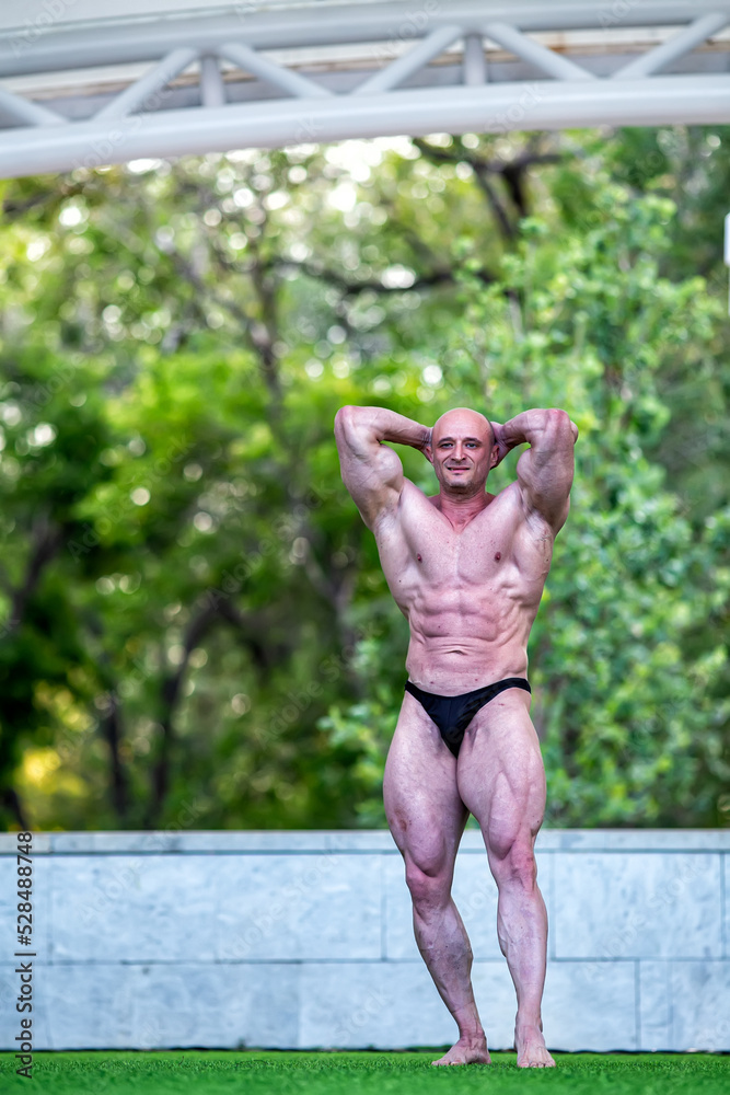 White man on stage with green grass demonstrates a beautiful male body with tensed muscles