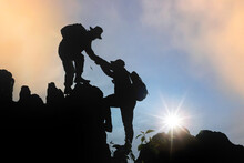 Silhouette Of Hiker Helping Each Other Hike Up A Mountain At Sunset.