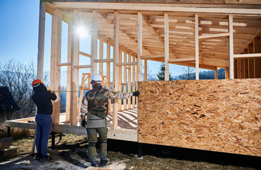 Canvas Print - Men workers building wooden frame house on pile foundation. Back view of carpenters using tape measure for measuring wooden planks. Carpentry concept.