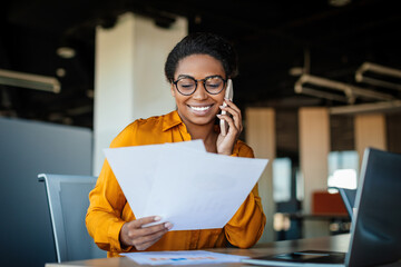 Happy black businesswoman talking on cellphone and checking documents in office, working with papers at workplace