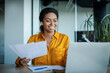 © Prostock-studio - Happy black businesswoman working with papers and having video call on laptop, sitting in office