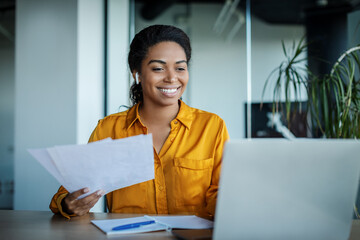 Happy black businesswoman working with papers and having video call on laptop, sitting in office