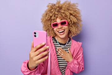 Wall Mural - positive curly haired young european woman wears trendy sunglasses striped jumper and jacket takes s