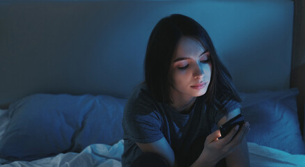 Online communication. Sleepless night. Pretty smiling woman sitting o bed scrolling smartphone in light shadow room interior.