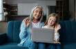 © Prostock-studio - Happy european small girl and mature woman look at laptop, waving hand in living room interior