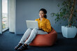 © Prostock-studio - Positive black female manager working on laptop computer in office, sitting on pouf beanbag chair, free space