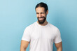 © khosrork - Portrait of cheerful man with beard wearing white T-shirt being in good mood, smiling broadly and winking at camera with toothy smile. Indoor studio shot isolated on blue background.