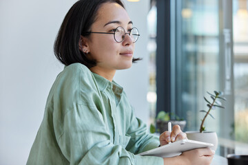 Talented female designer works with digital touch pad and holds stylus wears spectacles and shirt prepares project focused forward with thoughtful expression. People distance job and technology