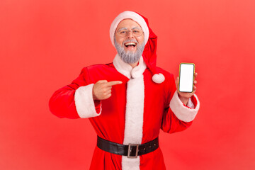 Wall Mural - portrait of excited elderly man with gray beard in santa claus costume standing with smart phone in 