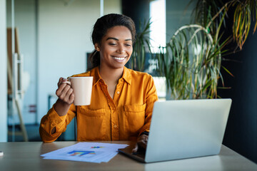 Aufkleber - Happy african american businesswoman working on laptop and drinking coffee, sitting at workplace in office
