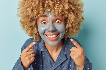 Wall Mural - Horizontal shot of positive woman with curly hair smiles toothily shows white teeth applies facial clay mask for rejuvenation dressed in slumber suit isolated over blue background. Skin care concept