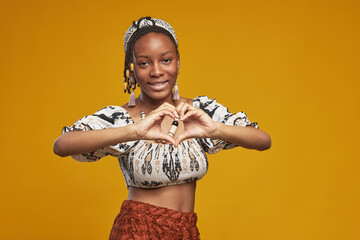 Portrait of African teenage girl making heart shape with her hands and smiling at camera standing against yellow background