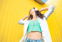 A Full-length View Of A Beautiful Cheerful Girl Jumping While Listening To A Melody Isolated Against A Bright Yellow Background. A Young Woman With Headphones Dancing On The Street . Generation Z
