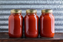 Three Jars Of Homemade Water Bath Canned Crushed Tomato Sauce Made From Homegrown Tomatoes On A Wooden Table And Industrial Background