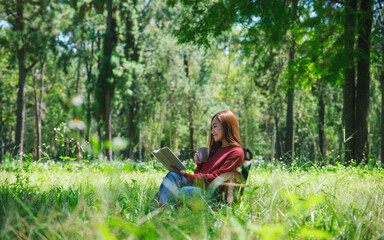 Wall Mural - Portrait of a beautiful young asian woman reading a book and drinking coffee while sitting on a camping chair in the park