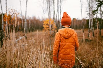 Wall Mural - Thoughtful ten years girl in orange coat stands in fall forest among high dry grass and golden trees. Autumn vibes, outdoor lifestyle. Hello autumn, thinking and mental health. View from back