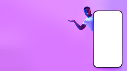 Poster - Happy young black man offering blank space from behind big cellphone with empty white screen in neon light, mockup