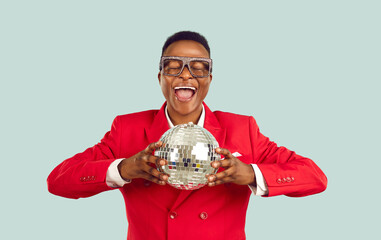 Wall Mural - Young African American man entertainer in stylish glasses and red suit clutches disco ball symbolizing relaxation in nightclub and fun rave standing on pastel blue background. Party producer portrait