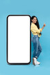 © Prostock-studio - Lovely young woman standing near big mobile phone with empty white screen and showing thumb up gesture, mockup