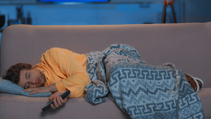 young man in yellow hoodie holding remote controller and lying under blanket on sofa