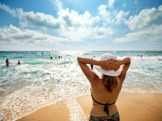 Tanned girl with a white hat, relaxes on the beach looking at the sea on a sunny summer day, in the background bathers cool off by the sea in the Gulf of Policastro, Italy.