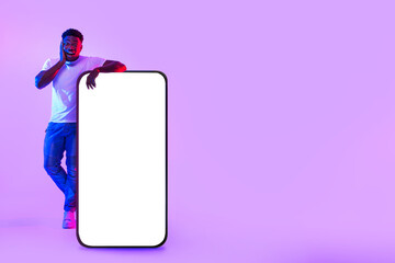 Poster - Surprised young black man standing near giant cellphone with empty white screen in neon light, mockup for mobile app