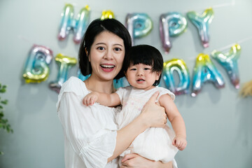 Wall Mural - front portrait of a beautiful asian mother smiling at the camera with her baby daughter in arms at a blurred background with birthday decoration.