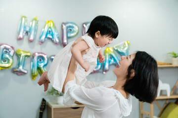 Wall Mural - side view of a cheerful Korean mother holding her lovely baby daughter high up with the wall decorated for birthday at background.