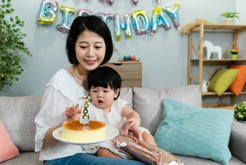 Wall Mural - pretty smiling japanese mother is holding and looking at the cake in hands while her cute baby girl reaching for the candle with curiosity at a birthday party.