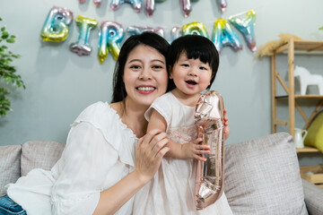 Wall Mural - portrait of a happy chinese mom holding her cute baby child and looking at the camera with smile in a birthday party at home