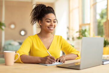 Wall Mural - Smiling african american lady working on laptop taking notes in notebook, sitting at desk at home, copy space