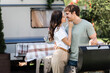 © LIGHTFIELD STUDIOS - Side view of woman kissing boyfriend near grill and camper van