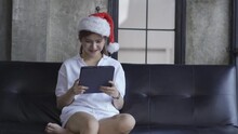 Asian Woman In White T-shirt Wearing Red Santa Hat Playing Tablet On Sofa At Home
