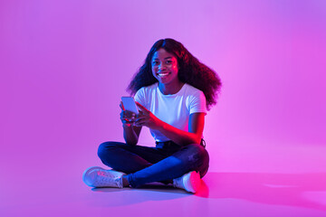 Poster - Young African American female sitting cross legged with mobie phone, surfing internet, chatting online in neon light