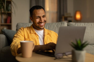 Poster - Smiling young black man working or communicating online, sitting on couch with laptop at home