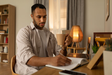 Poster - Serious young black man sitting at desk, working on laptop, taking notes during remote lesson, drinking coffee at home