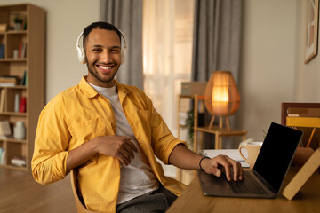 Wall Mural - Young happy black man in headphones using laptop pc with mockup, working online at home, copy space