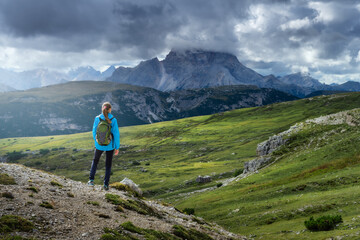 Wall Mural - Girl with backpack on the hill looking at mountains at sunset in summer. Dramatic landscape with young woman, high rocks, trail, green grass, cloudy sky in Dolomites, Italy. Adventure and hiking