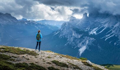Wall Mural - Girl with backpack on the hill looking at mountains at sunset in summer. Dramatic landscape with young woman, high rocks, trail, green grass, cloudy sky in Dolomites, Italy. Adventure and hiking
