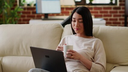 Poster - Happy asian woman sipping beverage while working remotely on laptop at home. Smiling heartily young adult person enjoying remote work while sitting on sofa inside modern apartment.