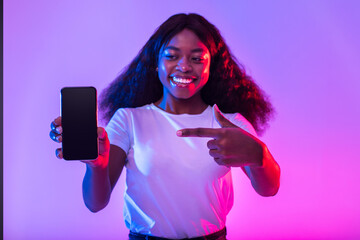 Poster - Smiling millennial black female pointing at cellphone with empty screen in neon light, mockup for website or mobile app
