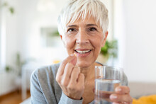 Senior Woman At Home Sitting At Table And Kitchen At Home Holding Cup Of Water And Pill Coronavirus Treatment Close-up Blurred Background