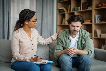 Wall Mural - Serious young black female doctor calms european guy, client suffering from depression in office clinic interior